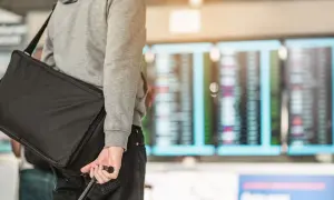 Mann mit schwarzer Reisetasche und Koffer vor Anzeigetafel am Flughafen.